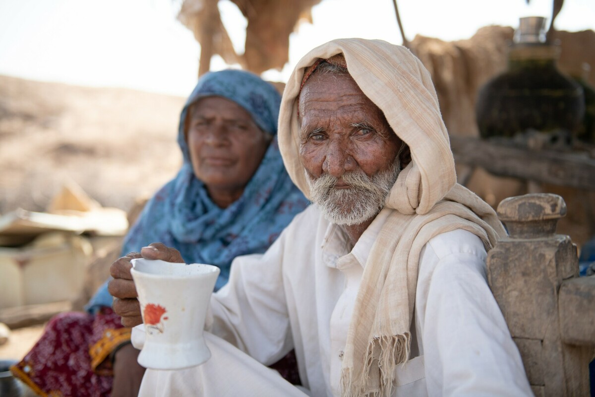 This image shows an older man and woman in the desert in Pakistan. The woman is wearing a blue head scarf, the man is holding a cup in his hand, and they both are looking at the camera in a serious way.
